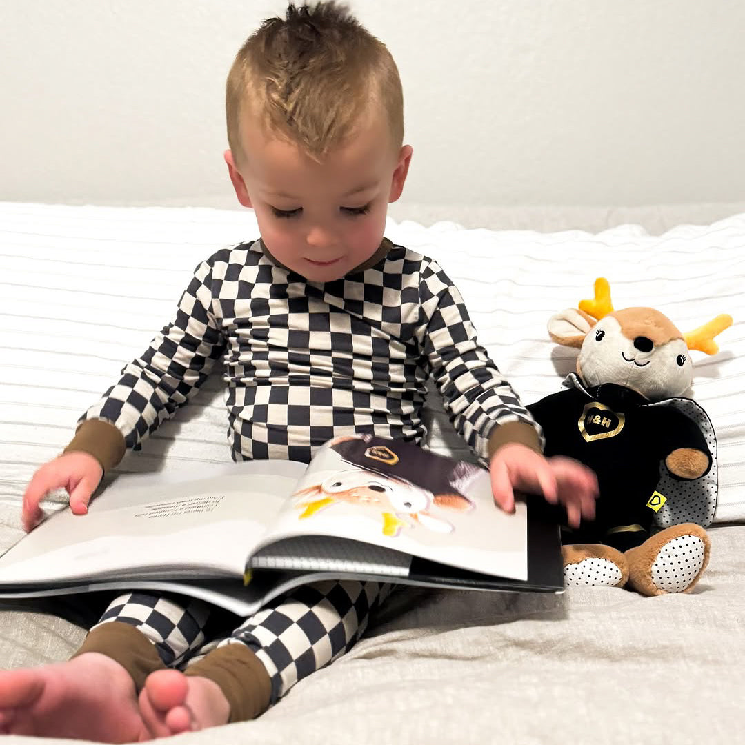 Child in checkered pajamas reading a book with a plush Hartie doll on a bed.
